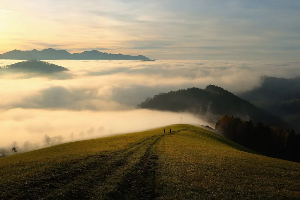 Weg über einen begrasten Höhenzug, im Tal darunter liegt eine Nebeldecke, dahinter Berge im Morgenlicht